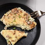 A fork holds a piece of a fluffy omelet served on a black plate, reminiscent of a savory French toast casserole. The omelet contains chunks of meat and is topped with chopped green onions, similar to crispy turkey bacon. The rest reveals a soft, moist interior that's partly eaten.