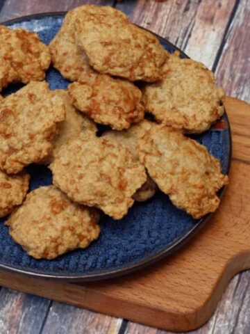A blue plate filled with several round, baked cookies is placed on a wooden serving board. The surface beneath is textured wood in shades of brown and gray. The cookies appear golden brown and slightly irregular in shape.