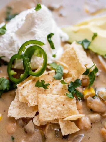 A bowl of creamy soup topped with crushed tortilla chips, sliced jalapeño, chopped cilantro, and a dollop of sour cream. Avocado slices are on the side. The soup contains ingredients like corn and beans.
