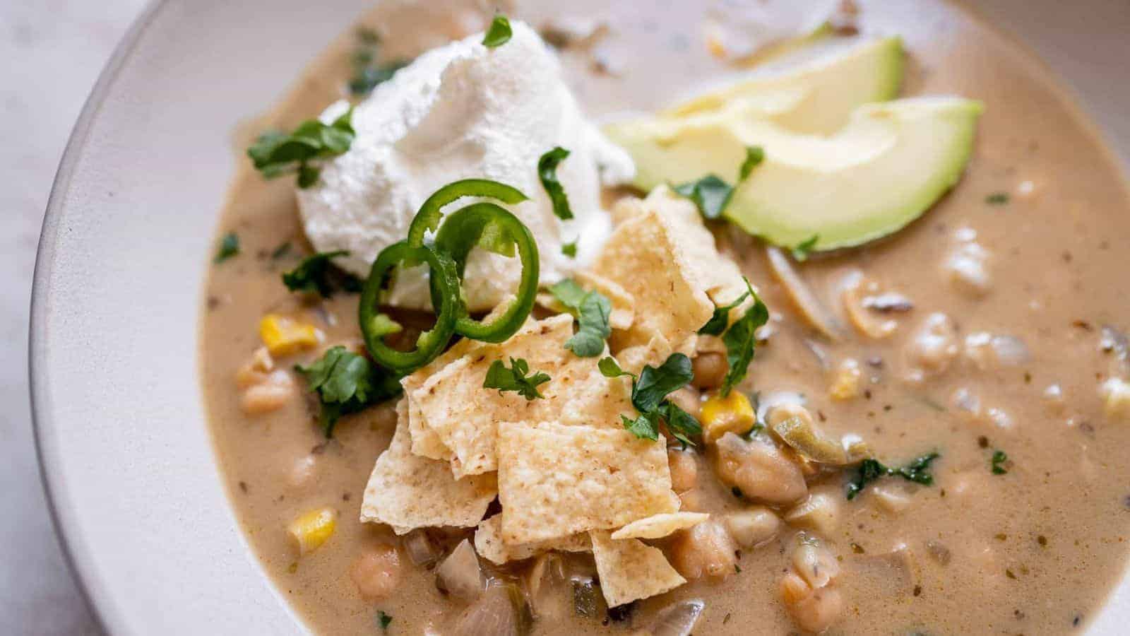 A bowl of creamy soup topped with crushed tortilla chips, sliced jalape&ntilde;o, chopped cilantro, and a dollop of sour cream. Avocado slices are on the side. The soup contains ingredients like corn and beans.