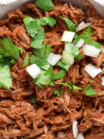 A bowl of shredded meat garnished with chopped cilantro and diced onions. The meat appears to be seasoned and cooked in a sauce. The bowl is placed on a wooden surface with cilantro leaves nearby.