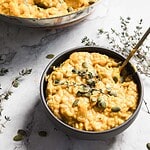 A bowl of creamy pumpkin risotto, infused with tender chicken pieces and garnished with pumpkin seeds and a sprig of fresh thyme. The dish rests on a marble surface, surrounded by additional sprigs of thyme. A spoon is tucked invitingly in the bowl.
