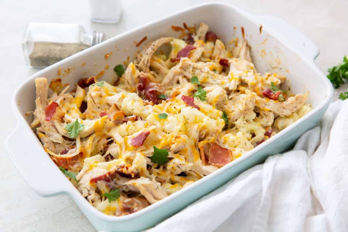 A white baking dish brimming with a delicious casserole of shredded chicken, crispy bacon pieces, and melted cheese, all sprinkled with chopped parsley. Ideal for chilly nights, this hearty dish sits next to a pepper shaker and white cloth on a light surface.