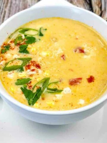A bowl of creamy yellow soup garnished with chopped green onions, cilantro, and paprika. The bowl is placed on a white square plate with a spoon and lime wedges on the side, set on a wooden table.