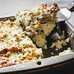 A spatula lifts a piece of baked casserole from a metal baking dish, revealing layers reminiscent of Texas French Toast. The dish features a browned, cheesy top, visible herbs, and a dense filling that's irresistible.