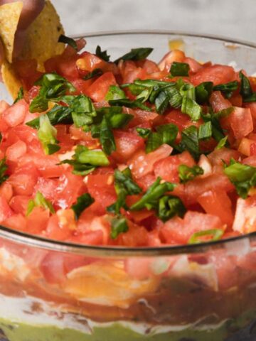A hand dips a tortilla chip into a layered dip in a glass bowl. The dip consists of chopped tomatoes, green onions, cheese, beans, guacamole, and sour cream, arranged in visible layers. The bowl is on a marble surface, with more chips in the background.