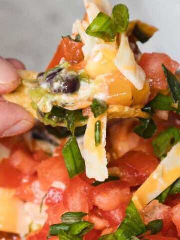 A hand holds a chip topped with cheese, beans, green onions, and diced tomatoes above a bowl of dip—perfect for sharing at any outdoor party recipes gathering.