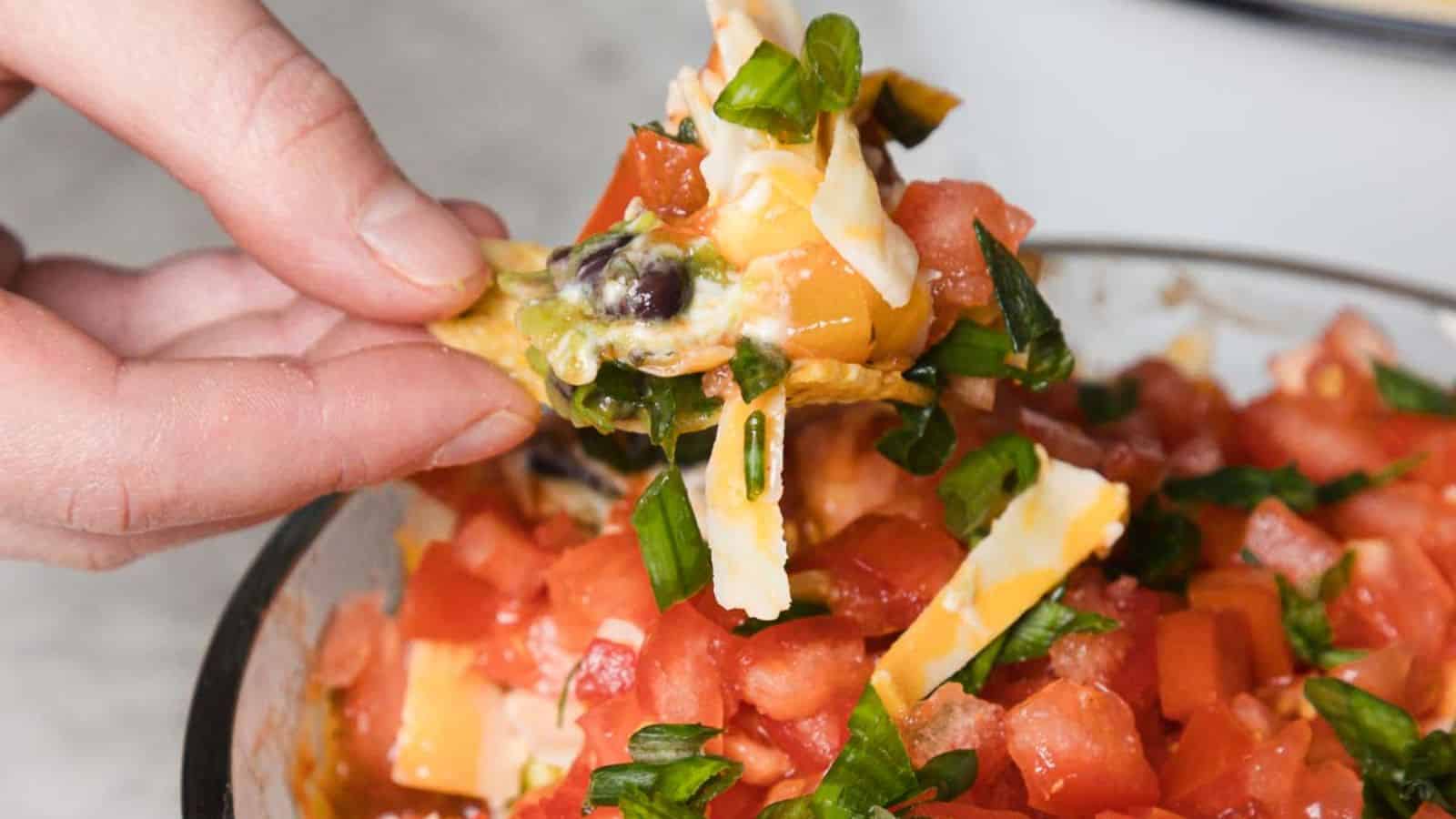 A hand dips a tortilla chip into a layered dip in a glass bowl. The dip consists of chopped tomatoes, green onions, cheese, beans, guacamole, and sour cream, arranged in visible layers. The bowl is on a marble surface, with more chips in the background.