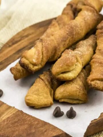Twisted pastry sticks sprinkled with sugar are placed on a wooden serving board lined with parchment paper. A few chocolate chips are scattered on one side. In the background, there's a small wooden bowl with a dark spread.