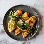 Grilled chicken breasts on a dark plate garnished with chopped herbs and two lemon wedges. The plate is on a marble surface. Reminiscent of an air fryer recipe, the chicken is seasoned with visible spices for that extra burst of flavor.