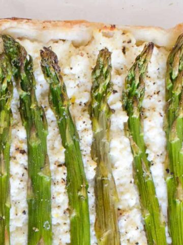 A rectangular tart topped with whole asparagus spears and a layer of ricotta cheese on a pastry crust. The edges are slightly browned, and the tart is on a parchment-lined surface.