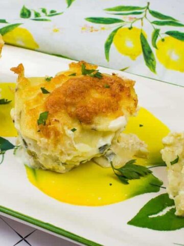 Three servings of potato and cheese gratin are presented on a rectangular plate with a lemon and leaf pattern. The gratin is topped with a golden brown crust and garnished with fresh herbs. A lemon-themed cloth is in the background.