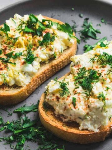 Two slices of bread topped with egg salad and garnished with chopped herbs are placed on a dark gray plate. Additional herbs and lemon slices are visible in the background on a light surface.