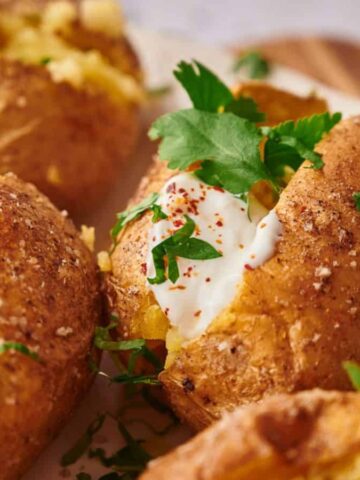 Baked potatoes with crispy skins are topped with sour cream, chopped cilantro, and a sprinkle of paprika, creating a hearty comfort meal reminiscent of Grandma’s magic. They are beautifully arranged on a white plate.