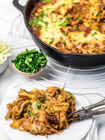 A plate of baked pasta with melted cheese, served with a fork and spoon. In the background, a skillet filled with more pasta sits on a cooling rack. Small bowls of shredded cheese and chopped herbs are adjacent to the skillet.