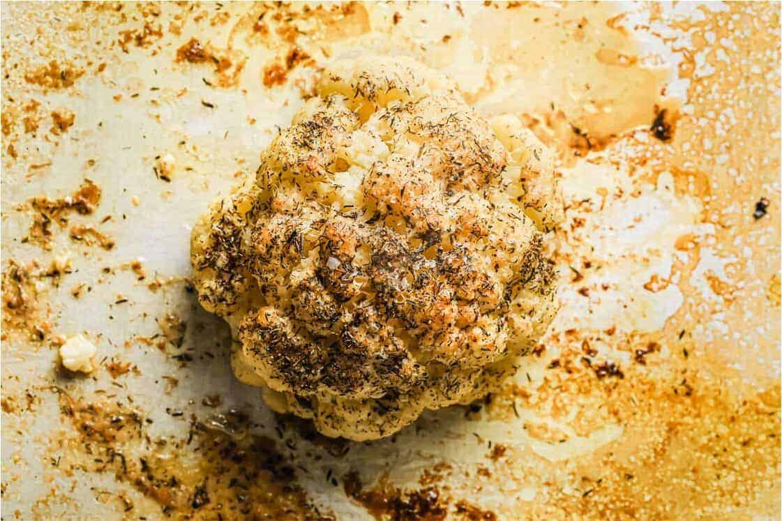 Whole roasted cauliflower, seasoned with herbs, rests on a baking sheet amidst browned oil and seasoning remnants&mdash;a cozy recipe perfect for those nights when procrastination is the main course.