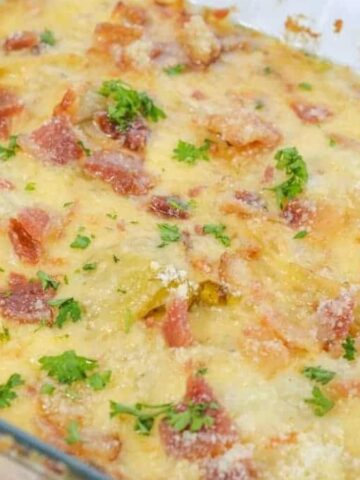 A glass baking dish filled with a creamy, baked casserole. The top is golden brown with visible pieces of crispy bacon, melted cheese, and sprinkled parsley. Edges show slight browning, indicating a well-cooked dish.