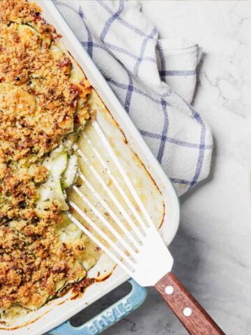 A baked dish with a golden-brown crumb topping is in a rectangular baking dish, which has a white and blue checkered cloth nearby. A serving is on a round plate to the right. The surface is a light marble texture.