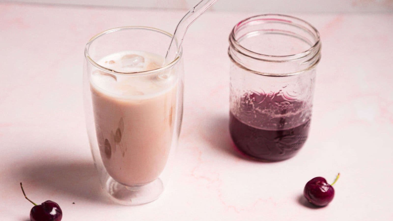 A glass of pink iced beverage with a clear straw next to a jar containing a dark purple liquid. Two cherries lie beside them on a light pink surface.