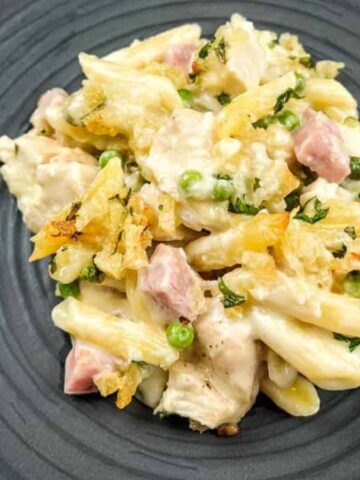 A black plate with a serving of creamy pasta bake featuring penne, diced ham, green peas, and melted cheese. A fork and knife are placed beside the plate on a wooden surface.