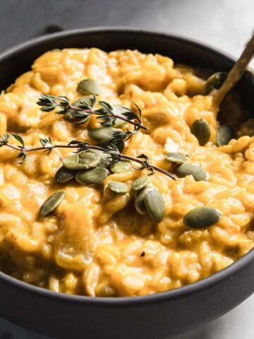 A black bowl filled with creamy pumpkin oatmeal garnished with fresh thyme and pumpkin seeds. A spoon rests inside the bowl. The background is a light gray marble surface scattered with a few pumpkin seeds.