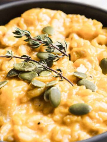 A bowl filled with creamy orange-yellow risotto, topped with pumpkin seeds and sprigs of fresh thyme. A spoon is placed in the risotto. The background is blurred, featuring additional thyme sprigs.