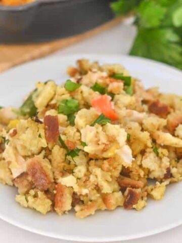 A plate of stuffing with visible chunks of bread, herbs, and vegetables sits on a white surface. A bunch of fresh parsley peeks from the background alongside a portion of a pan, adding to the inviting ambiance of hearty family-size casseroles perfect for cherished family meals.