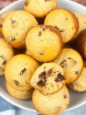 A bowl filled with small chocolate chip muffins. One muffin has a bite taken out, revealing the chocolate chips inside. A blue and white checkered cloth is partially visible on the side.