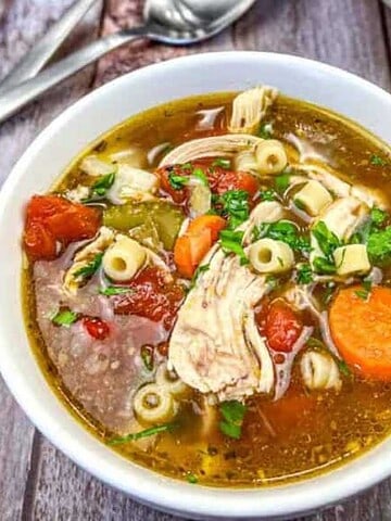 A white bowl filled with comforting soup sits on a wooden table, featuring pasta, chunks of chicken, tomato pieces, sliced carrots, and green herbs. This delightful comfort food is a haven for your taste buds. Silverware and a white napkin are partially visible in the background.