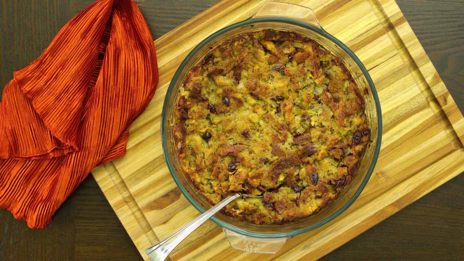 A round glass baking dish filled with baked stuffing sits on a wooden cutting board. A metal spoon rests inside the dish. An orange cloth is draped to the left of the dish. Brown bits are visible on the surface of the stuffing.
