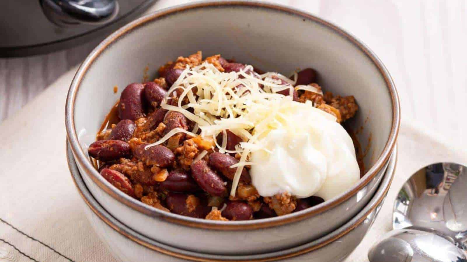 A bowl of Budget Comfort Foods chili, topped with shredded cheese and a dollop of sour cream, warms your heart. It features red kidney beans and ground meat in a gray ceramic bowl on a beige cloth, with two spoons beside it.