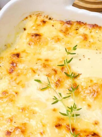 A baked potato gratin with a golden-brown, cheesy crust is in a white rectangular dish. Fresh thyme sprigs garnish the top. A wooden pepper mill is in the background, partially on a checkered cloth.