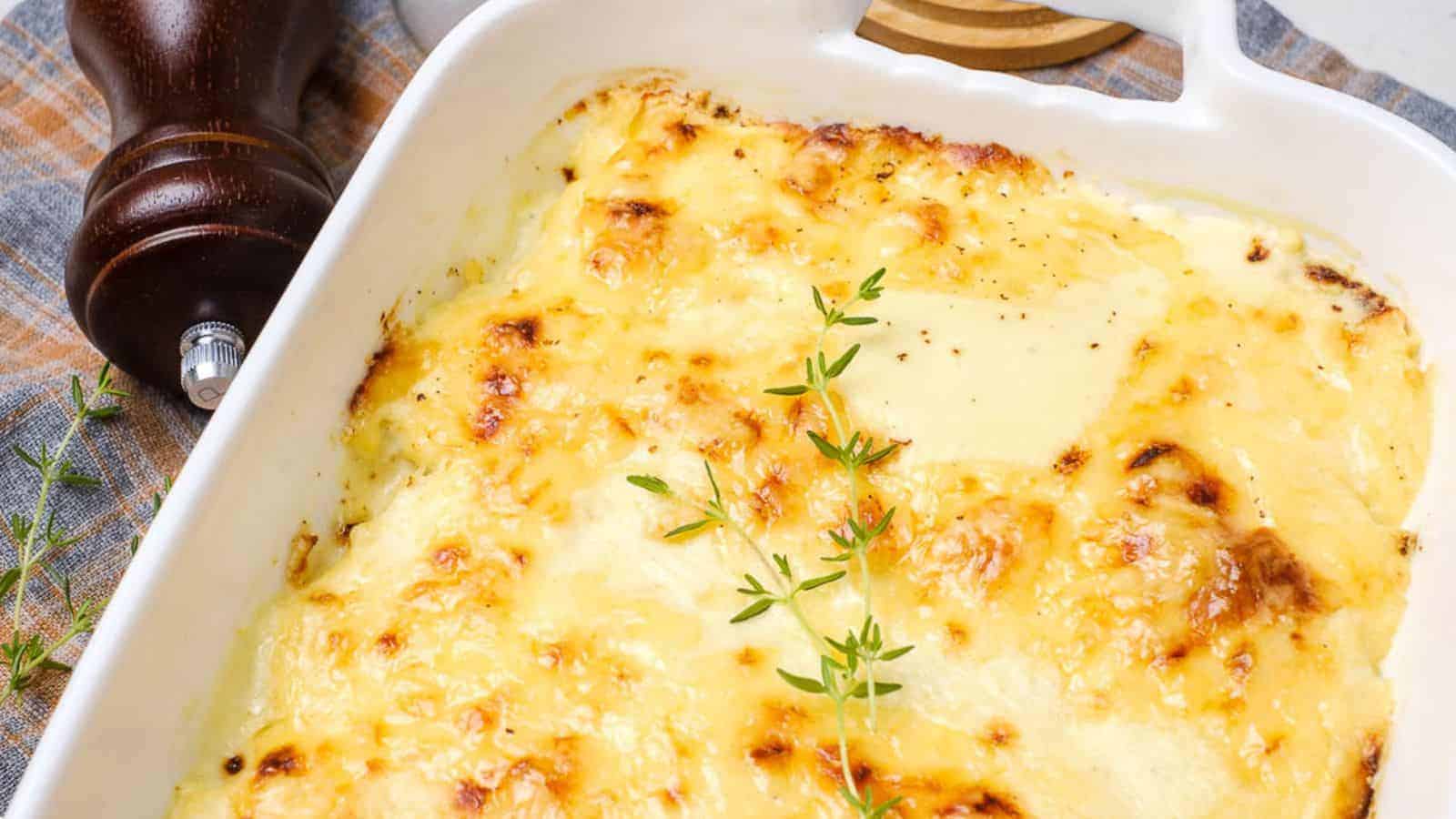 A baked potato gratin with a golden-brown, cheesy crust is in a white rectangular dish. Fresh thyme sprigs garnish the top. A wooden pepper mill is in the background, partially on a checkered cloth.
