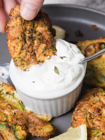 A hand dips a zucchini fritter into a white sauce in a small ramekin. The plate contains more fritters and lemon wedges. A spoon rests in the ramekin. The background features a marble surface.