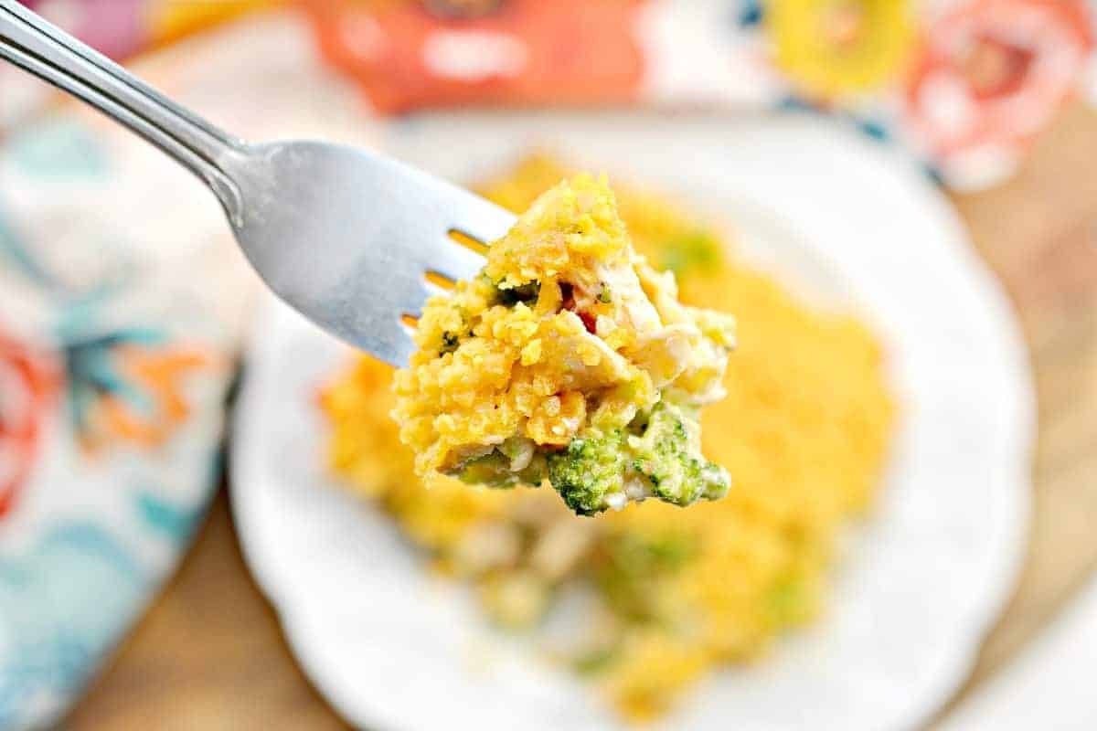A fork holds a bite of cheesy broccoli and rice casserole, with melted cheese and small broccoli pieces visible. A plate with more casserole is blurred in the background, set on a colorful tablecloth with floral patterns.