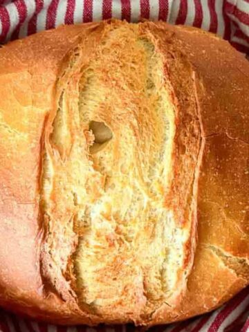 A round loaf of bread with a crusty brown exterior is placed on a red and white striped cloth. The bread is positioned centrally, and the cloth is gathered around its sides.