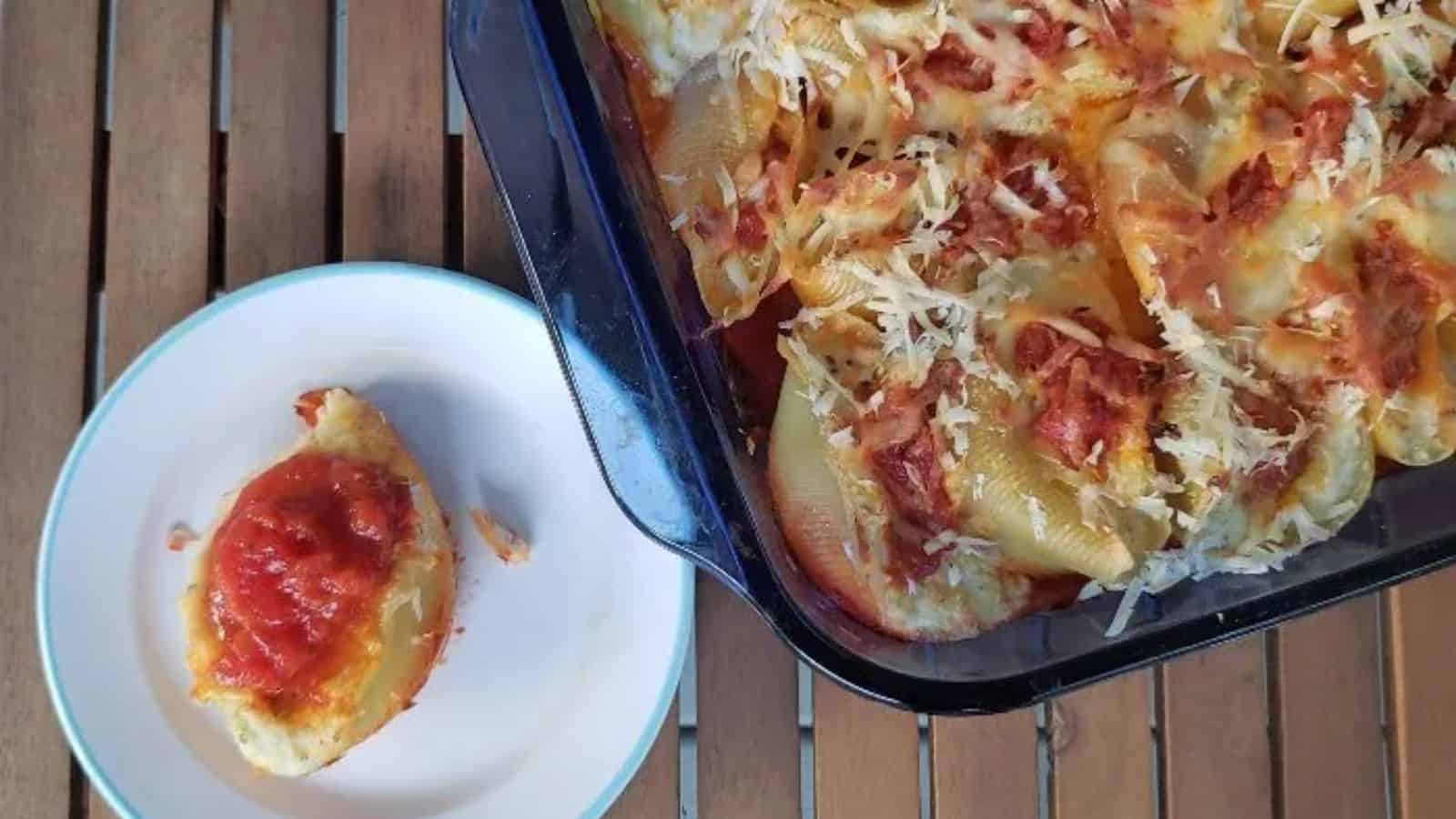 A baking dish filled with stuffed pasta shells topped with melted cheese and tomato sauce sits on a wooden table. A single stuffed shell, similarly topped, is served on a white plate with a green rim beside the dish.