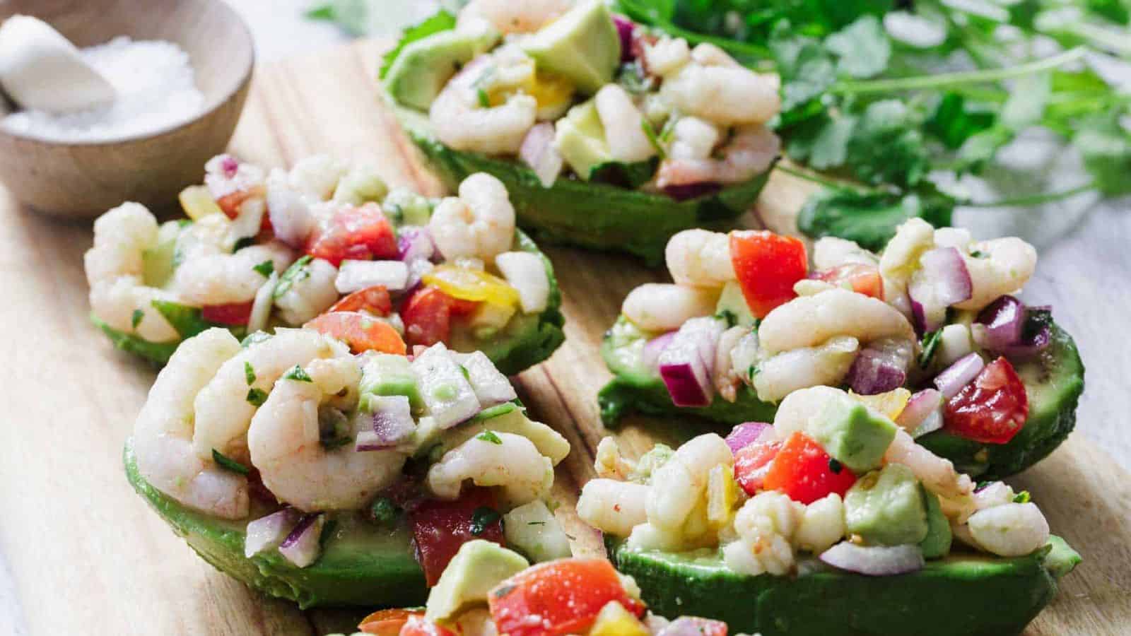 A black plate with a halved avocado filled with a shrimp salad, garnished with lime. A fork rests beside it. A wooden board holds more avocado halves in the background. A wooden bowl with mixed salad ingredients is partially visible.