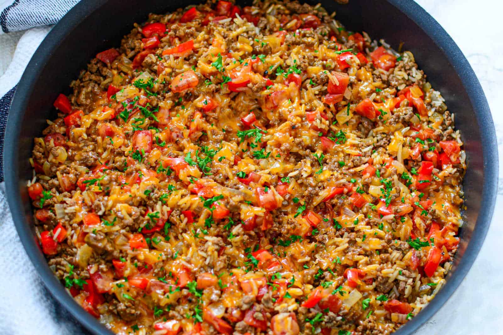 A skillet filled with a cooked dish of ground beef, rice, diced tomatoes, melted cheese, and garnished with chopped parsley. The mixture appears colorful with a blend of red, brown, and green.