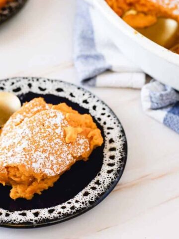 A slice of orange dessert topped with powdered sugar sits on a black and white plate with a spoon. In the background, a serving dish with more dessert is partially visible beside a white and blue towel.