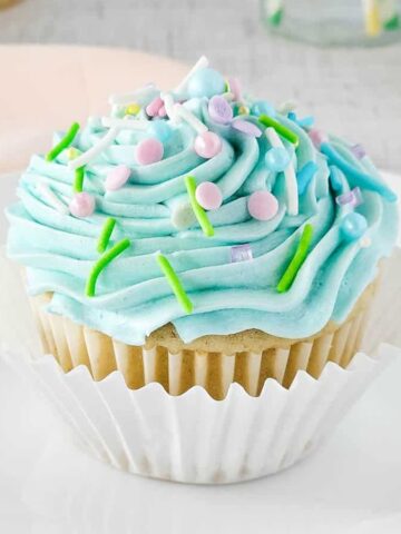 A cupcake with light blue frosting, decorated with green and pink sprinkles, is placed on a white plate. It is wrapped in a white paper liner and sits on a white-textured surface beside a light pink cloth.