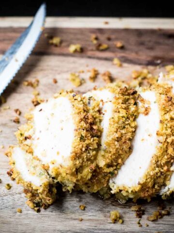 Sliced breaded chicken on a wooden cutting board, accompanied by a serrated knife. The chicken is coated with a crumb mixture and appears golden brown and crispy.