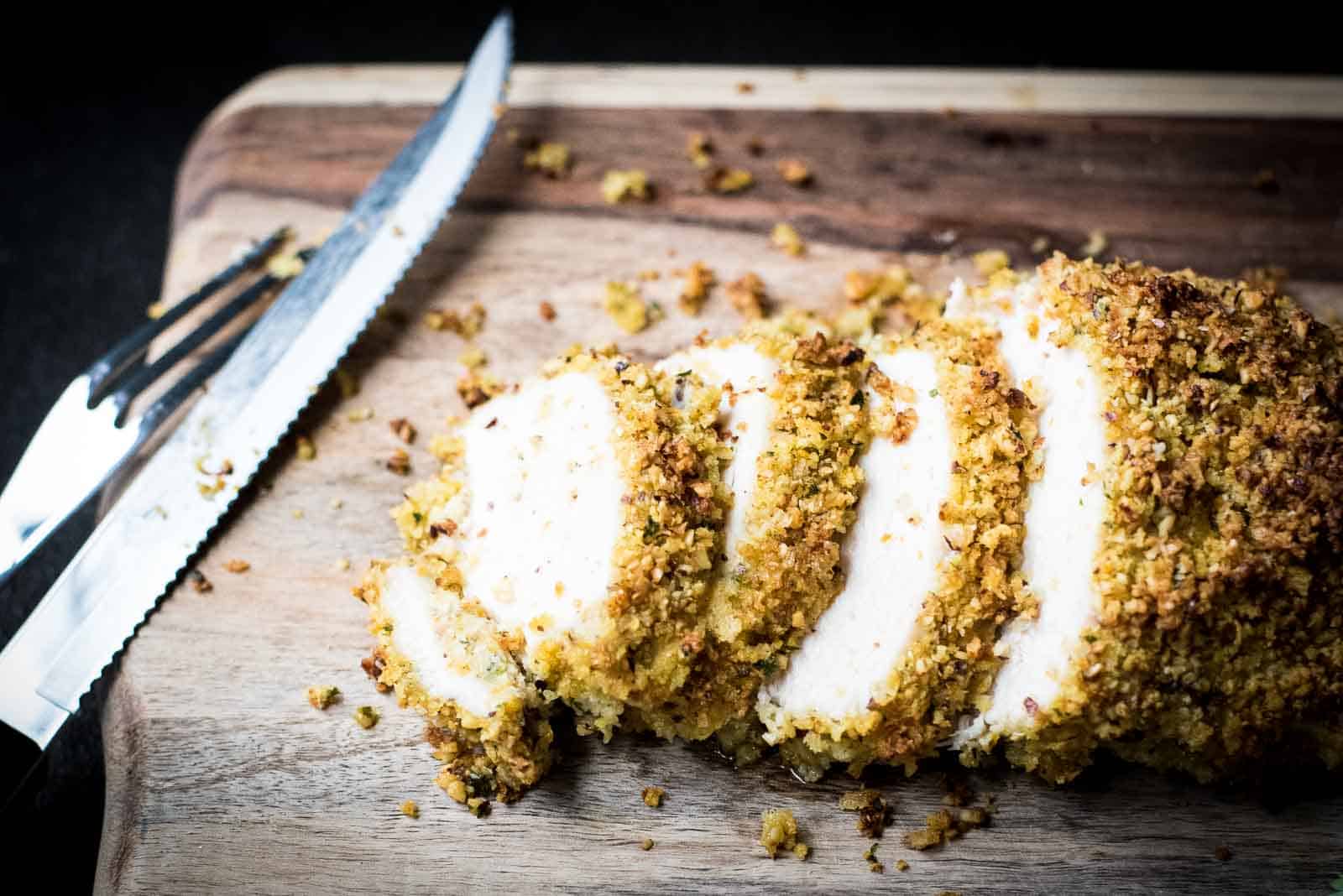 Sliced breaded chicken on a wooden cutting board, accompanied by a serrated knife. The chicken is coated with a crumb mixture and appears golden brown and crispy.