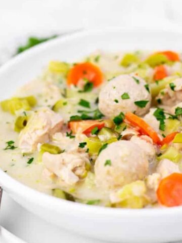 A white bowl filled with soup containing chicken pieces, sliced carrots, celery, and dumplings. The soup is topped with chopped parsley. In the background, there's a plate with assorted bread rolls.