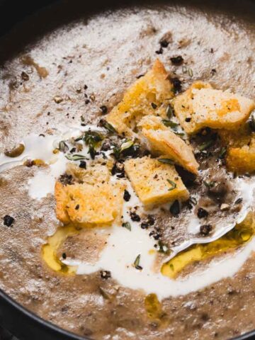 A bowl of creamy mushroom soup garnished with croutons, a drizzle of olive oil, and fresh herbs. A golden spoon lies nearby, and sprigs of thyme are placed on the table beside the bowl.
