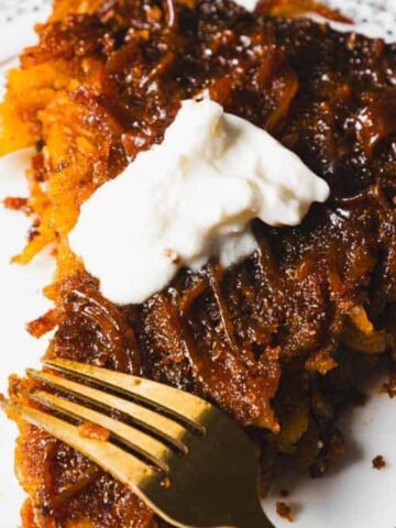 A slice of brown, crispy hash brown pie topped with a dollop of sour cream sits on a decorative black and white plate. A gold fork is partially inserted into the pie on the left side.