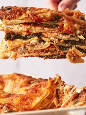 A hand holds a slice of vegetable lasagna above a dish. The lasagna layers include visible noodles, vegetables, and a cheese topping. The dish is rectangular, with the rest of the lasagna remaining inside.