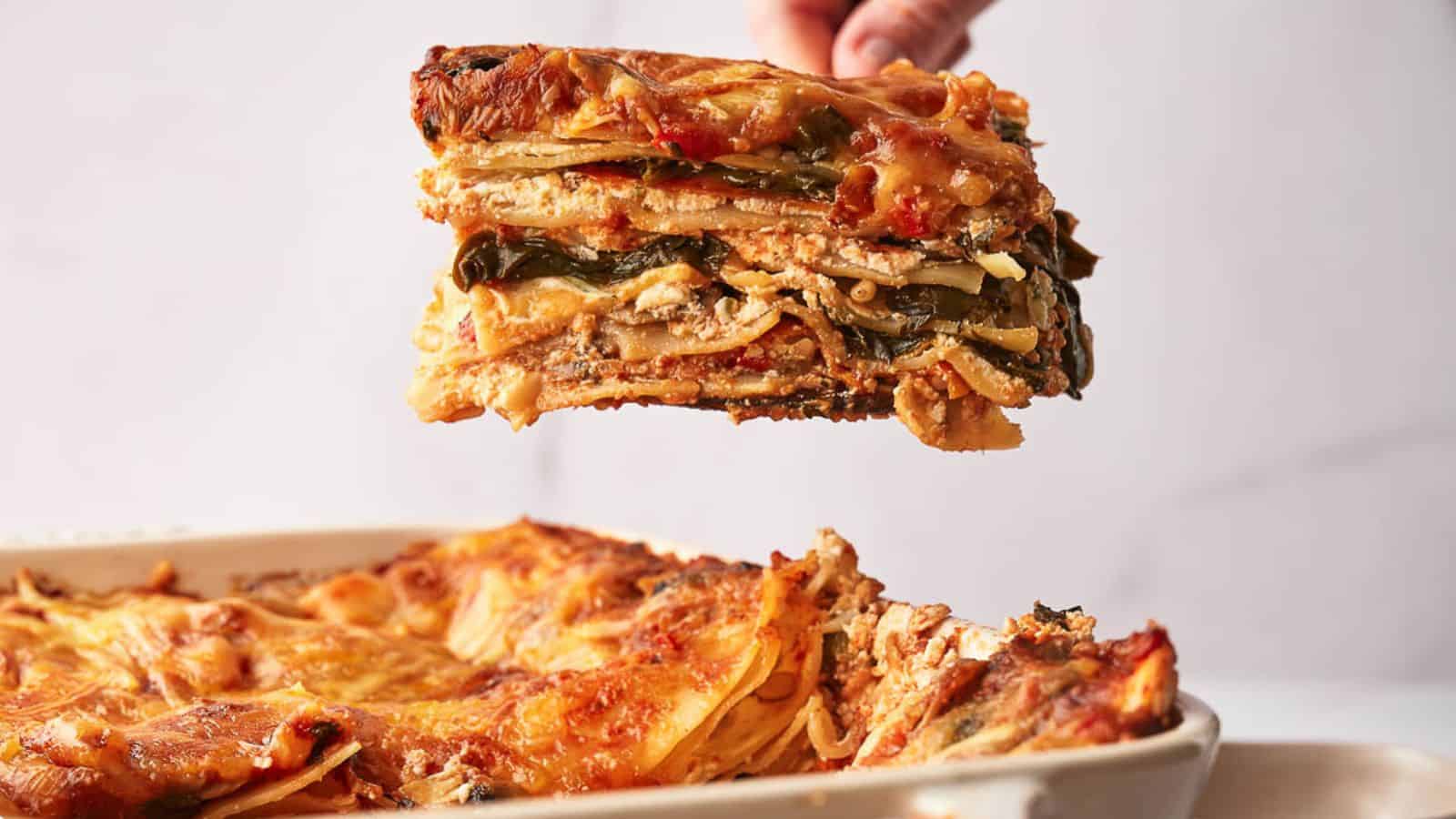A hand holds a slice of vegetable lasagna above a dish. The lasagna layers include visible noodles, vegetables, and a cheese topping. The dish is rectangular, with the rest of the lasagna remaining inside.