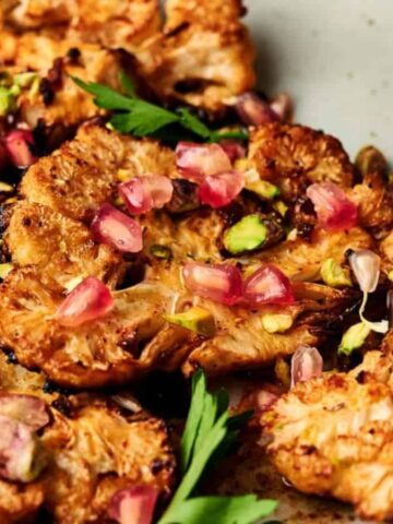 Grilled cauliflower steaks on a plate, topped with pomegranate seeds, chopped pistachios, and garnished with fresh parsley. The dish has a golden-brown, crispy appearance.