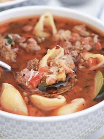 A bowl of soup containing chunks of meat, shell pasta, zucchini slices, and herbs in a tomato-based broth. A spoon rests in the bowl. A whole tomato, celery stalk, and a striped cloth are partially visible in the background—true budget comfort food to warm your heart.